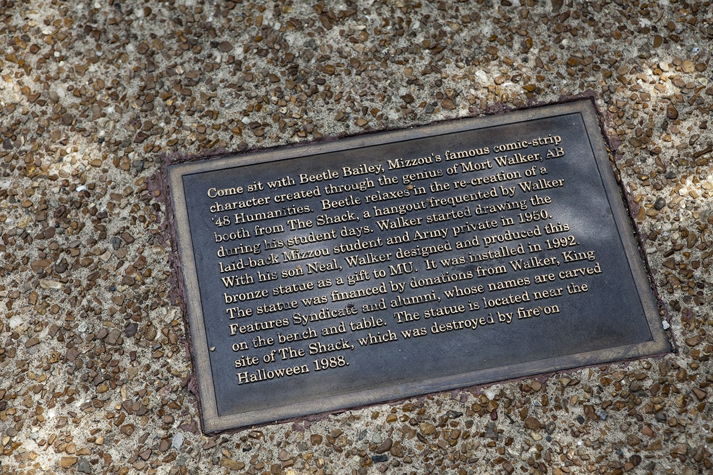 "Come sit with Beetle Bailey, Mizzou's famous comic-strip character created through the genius of Mort Walker, AB '48 Humanities.  Beetle relaxes in the re-creation of a booth from The Shack, a hangout frequented by Walker during his student days.  Walker started drawing the laid-back Mizzou student and Army private in 1950.  With his son Neal, Walker designed and produced this bronze statue as a gift to MU.  It was installed in 1992.  The statue was financed by donations from Walker, King Features Syndicate and alumni, whose names are carved on the bench and table.  The statue is located near the site of The Shack, which was destroyed by fire on Halloween 1988."