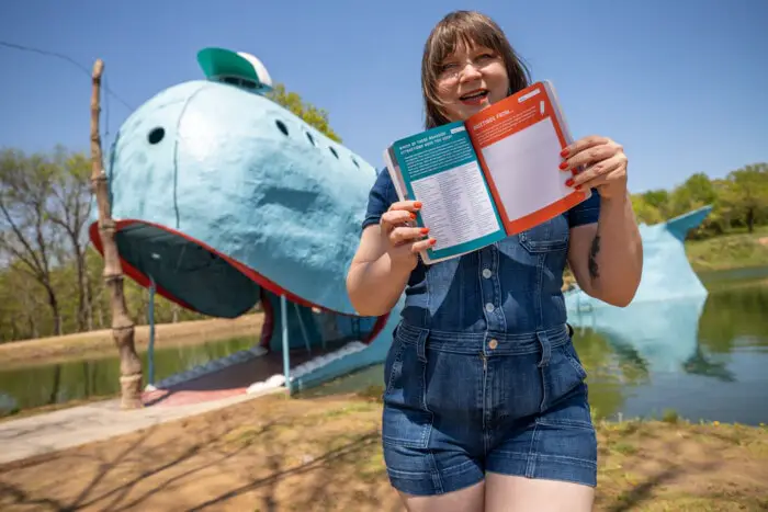 The Road Trip Journal & Activity Book - Which Roadside Attractions Have You Seen? Road Trip Game - Reading a road trip book in front of the Blue Whale of Catoosa in Oklahoma Route 66.