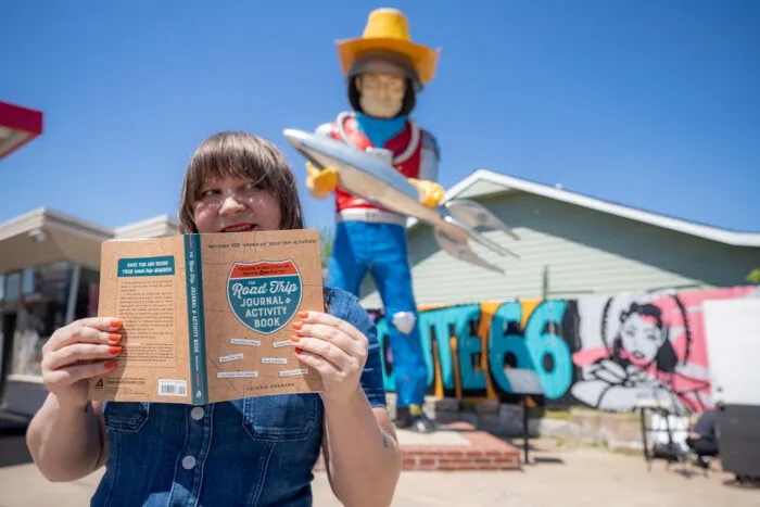 The Road Trip Journal & Activity Book - Reading a road trip book in front of the muffler man at Buck Atom's Cosmic Curios on Route 66 in Tulsa, Oklahoma